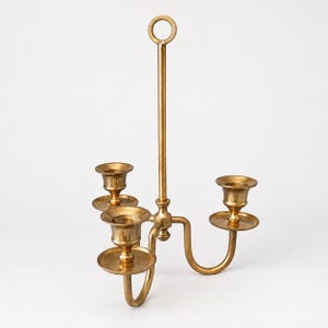 May include: A polished brass candelabra featuring three candle holders arranged around a central vertical stem. The stem is topped with a circular ring handle, and the three arms extend outward to support individual drip plates and candle cups.
