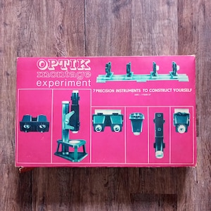 May include: A rectangular pink box for the Optik montage experiment kit. The box features white text reading Optik montage experiment and 7 precision instruments to construct yourself. It displays illustrations of various optical components.