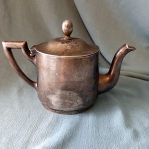 Op de afbeelding: Een doffe, zilverkleurige metalen theepot met een cilindrisch lichaam, een gebogen handvat, een korte tuit en een deksel met een kleine bolvormige knop. Het oppervlak vertoont zichtbare tekenen van oxidatie en slijtage met een donkere patina.