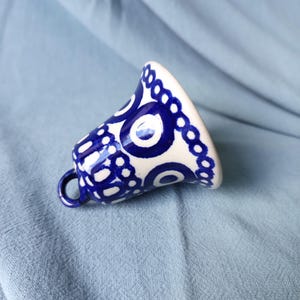 Op de afbeelding: Een keramische bel met een glanzende crèmekleurige afwerking en een handgeschilderd patroon van donkerblauwe cirkels en onderling verbonden ringen. De bel heeft een klein lusje aan de bovenkant om op te hangen en een uitlopende opening.