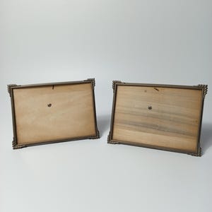 May include: Two rectangular picture frames shown from the back, featuring dark metallic borders with decorative corner detailing. The frames have light brown cardboard backing panels secured with small metal fasteners and a central metal pin.