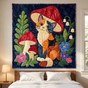 Op de afbeelding: Een vierkante stoffen wandhanger met een lapjeskat die onder een grote rode paddenstoelhoed zit. De scène bevat groene varenbladeren, twee kleinere paddenstoelen, witte lelietjes-van-dalen en blauwe en roze bloemen op een donkerblauwe achtergrond.