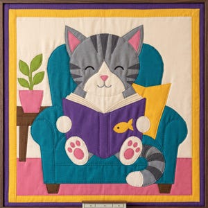 May include: A fabric wall hanging featuring a grey tabby cat with pink ears and paw pads sitting in a teal armchair. The cat holds an open purple book with a yellow fish icon. A small potted green plant sits on a brown side table nearby.