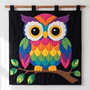 Könnte beinhalten: Ein rechteckiger Wandbehang aus Stoff mit einer bunten Eulen-Applikation auf schwarzem Hintergrund. Die Eule hat große gelbe Augen, einen gelben Schnabel und mehrfarbige Federn in den Farben Pink, Orange, Gelb, Grün, Blau und Violett.
