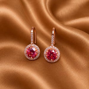 May include: A pair of rose gold drop earrings featuring a round, vibrant pink center stone surrounded by a halo of small, clear white crystals. The hook-style earring posts are also accented with a vertical row of tiny, clear white crystals.