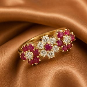 May include: A gold ring featuring three flower-shaped clusters of gemstones. The two outer flowers consist of red rubies surrounding a central white diamond, while the middle flower features white diamonds surrounding a central red ruby.