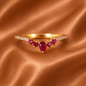 May include: A gold ring featuring a central pear-shaped red gemstone flanked by two smaller round red gemstones on each side, with a band accented by a row of small clear white stones extending along the curved V-shaped setting.