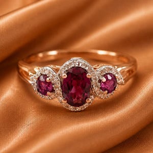 Puede incluir: Un anillo de oro rosa con una piedra preciosa roja profunda de corte ovalado en el centro, flanqueada por dos piedras rojas más pequeñas de corte redondo. Cada piedra está rodeada por un halo de pequeñas piedras blancas en una banda de oro rosa.