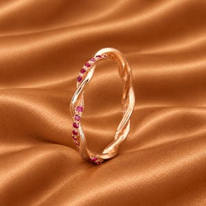 Könnte beinhalten: Ein Ring aus Roségold mit gedrehtem Banddesign. Eine Seite der Drehung ist glatt und poliert, während die andere Seite mit einer Reihe kleiner, runder, leuchtend rosaroter Edelsteine besetzt ist, die dem Verlauf des Metallbandes folgen.