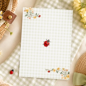 May include: A rectangular stationery sheet featuring a light green and white gingham pattern. The design includes a small red ladybug in the center and floral illustrations with yellow and orange blossoms in the top left and bottom right corners.