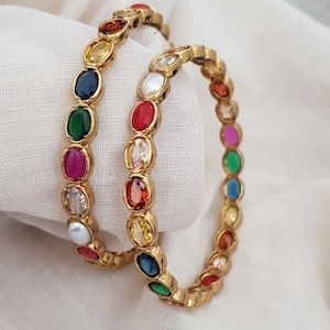 May include: Two gold-toned bangles featuring a series of oval-shaped, multi-colored gemstones set in a continuous row. The stones include shades of red, blue, green, yellow, white, and pink, creating a vibrant, repeating pattern around each band.