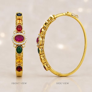 Puede incluir: Un brazalete rígido articulado de oro con una piedra preciosa roja ovalada central rodeada de pequeñas piedras blancas, flanqueada por otras piedras preciosas ovaladas rojas, verdes, amarillas y azules. El brazalete se muestra en vista frontal y lateral con etiquetas.