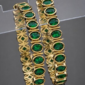 May include: Two gold-toned bangles featuring a series of oval-shaped, deep green faceted stones set in a continuous metal frame. The design showcases a repeating pattern of metallic links between each stone, creating a structured, circular jewellery piece.
