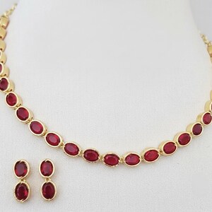 Puede incluir: Un collar de tono dorado y un par de pendientes a juego, cada uno con una serie de piedras facetadas de color rojo intenso en forma ovalada engastadas en metal pulido. El collar consiste en una fila continua de estas piedras rojas unidas.