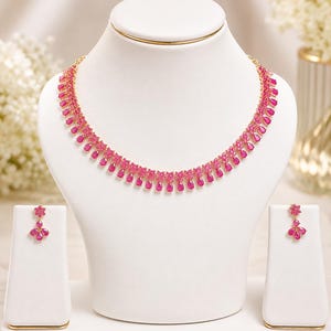 Puede incluir: Un conjunto de collar y pendientes en tono dorado con piedras de color rosa vibrante en forma de lágrima y pequeños detalles florales. El collar muestra una hilera continua de estas piedras rosas, mientras que cada pendiente tiene un diseño floral.