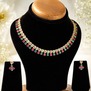 Puede incluir: Un conjunto de collar y pendientes en tono dorado con piedras rojas y verdes en forma de lágrima alternadas. El collar presenta un diseño de eslabones florales con pequeñas piedras claras, y cada pendiente muestra un grupo de tres piedras.