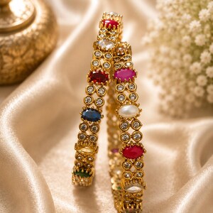 May include: Two gold-toned bangles featuring a series of small, clear, round stones set in a lattice pattern. Each bangle is accented with larger, oval-shaped, multi-colored stones in shades of red, blue, yellow, green, pink, and white.