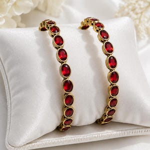 Peut inclure: Deux bracelets circulaires ornés d'une série de pierres facettées rouge foncé de forme ovale, serties dans une monture en métal doré. Les pierres sont disposées en ligne continue sur toute la circonférence de chaque bracelet, créant un motif uniforme.