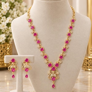 Puede incluir: Un conjunto de collar y pendientes en tono dorado con piedras redondas de color rosa vibrante. El collar presenta un diseño de colgante en forma de V con pequeñas piedras blancas, mientras que los pendientes reflejan este motivo floral.