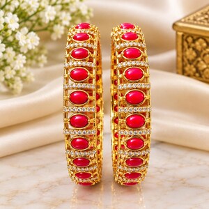 May include: Two gold-toned bangles featuring a repeating pattern of oval-shaped red stones set within a metal frame. Each bangle is accented with rows of small, clear, sparkling stones that line the edges and separate the red stone settings.