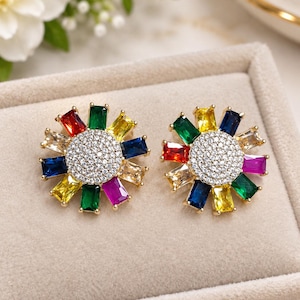 May include: A pair of gold-tone stud earrings featuring a central circular cluster of clear pavé stones surrounded by a sunburst arrangement of rectangular baguette-cut gemstones in vibrant shades of red, green, yellow, blue, and purple.