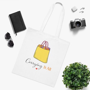 Puede incluir: Una bolsa de tela blanca con un gráfico de una bolsa de compras amarilla con asas rosas y el texto "Carrying It All" en letra cursiva negra junto a un pequeño corazón rojo. La bolsa cuenta con dos asas largas para llevar al hombro.