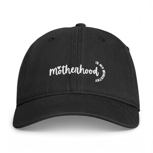 Puede incluir: Una gorra de béisbol negra con texto bordado en blanco que dice motherhood is my ministry, junto con un pequeño icono de corazón sobre la letra m. La gorra cuenta con una corona estructurada, una visera curva y ojales cosidos.