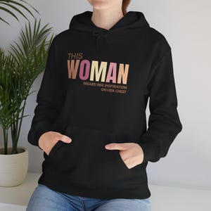 Puede incluir: Una sudadera negra con capucha que presenta el texto THIS WOMAN WEARS HER INSPIRATION ON HER CHEST impreso en un degradado de tonos marrón, beige, rosa y crema. La prenda incluye un bolsillo tipo canguro y una capucha con cordón.