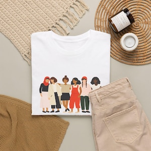 Può includere: Una t-shirt bianca a girocollo con un'illustrazione grafica di sei figure diverse in piedi una accanto all'altra. Le figure indossano vari abiti, tra cui un hijab rosso, un cappotto beige, una gonna nera, una salopette rossa e top con motivi decorativi.