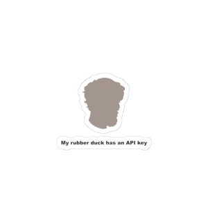May include: A sticker featuring a light brown silhouette of a person with curly hair and a beard, positioned above a white rectangular label with rounded corners that displays the black text, My rubber duck has an API key.