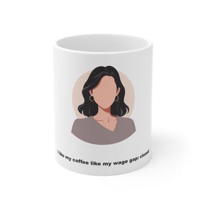 May include: A white ceramic mug featuring a minimalist illustration of a person with dark hair and hoop earrings against a circular tan background. Below the illustration, the text reads: I like my coffee like my wage gap: closed.