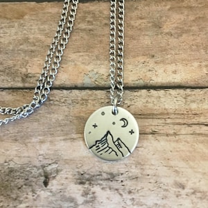 May include: A silver necklace with a round pendant featuring a hand-drawn design of mountains, a crescent moon, and stars.