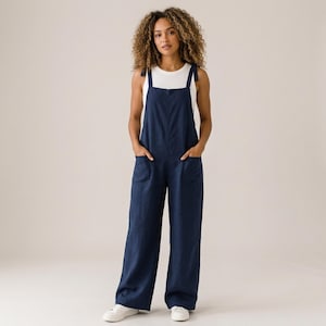 May include: A dark blue jumpsuit with wide legs and two front pockets, featuring shoulder straps that tie at the top. The jumpsuit is worn over a plain white sleeveless top. The garment has a relaxed fit and a solid colour throughout the fabric.