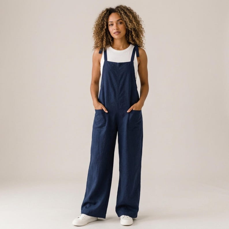 May include: A dark blue jumpsuit with wide legs and two front pockets, featuring shoulder straps that tie at the top. The jumpsuit is worn over a plain white sleeveless top. The garment has a relaxed fit and a solid colour throughout the fabric.