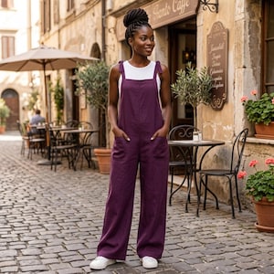 May include: A deep purple linen jumpsuit featuring wide legs, two front patch pockets, and shoulder straps tied in bows, worn over a simple white sleeveless top. The garment has a relaxed fit with a straight neckline and a solid, dark purple colour.