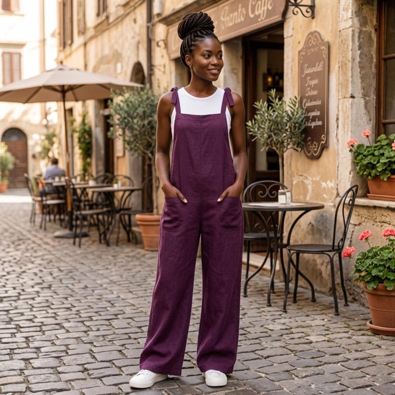 May include: A deep purple linen jumpsuit featuring wide legs, two front patch pockets, and shoulder straps tied in bows, worn over a simple white sleeveless top. The garment has a relaxed fit with a straight neckline and a solid, dark purple colour.
