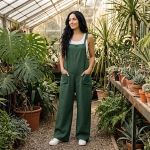 May include: A dark green sleeveless jumpsuit featuring a square neckline, wide-leg trousers, and two large front patch pockets. The garment is worn over a simple white tank top, creating a clean, functional silhouette with a relaxed fit.