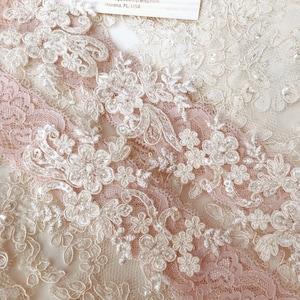 May include: Close-up of cream and blush pink lace fabric. The lace has detailed floral patterns with raised elements and small bead accents. The design incorporates multiple lace layers, producing a textured and elegant look. Ideal for bridal or craft projects.