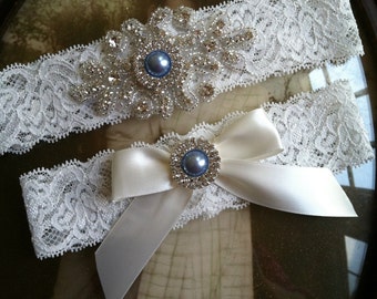 Pode incluir: Duas ligas de casamento de renda branca. A liga superior apresenta um grande adorno de prata e strass com um centro de pérola azul. A liga inferior tem um laço de cetim branco com um centro de pérola azul e strass semelhante.