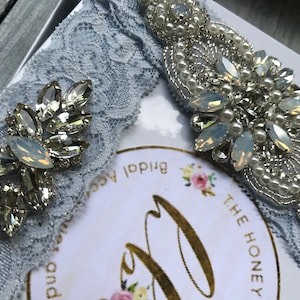 May include: A light blue bridal garter with two embellishments. One embellishment is a large cluster of rhinestones and pearls. The other embellishment is a smaller cluster of rhinestones. The garter is in a box with a label that says "The Honey Bridal Accessories".
