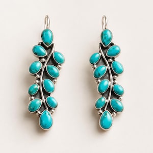 May include: A pair of silver dangle earrings featuring a branch-like design adorned with multiple oval-shaped turquoise stones. Each earring has a curved metal stem with small silver beads between the stones and a hook for attachment.