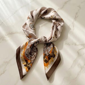 May include: A square scarf in a light cream color featuring a repeating pattern of brown feathers, orange leather-style belts, and small floral motifs. The scarf has a dark brown border and is tied in a loose knot, displaying a smooth, satin-like finish.