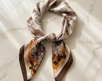 May include: A square scarf in a light cream colour featuring a repeating pattern of brown feathers, orange leather-style belts, and small floral motifs. The scarf has a dark brown border and is tied in a loose knot, displaying a smooth, satin-like finish.