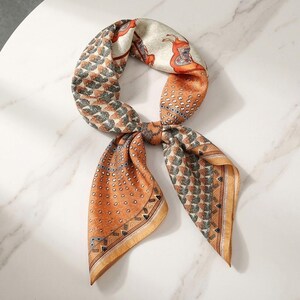 May include: A square scarf featuring a multi-colored pattern of orange, cream, and dark gray. The design includes stylized horse figures, geometric shapes, and dotted borders. The scarf is tied in a knot, displaying a mix of intricate prints.