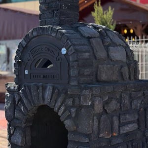Könnte beinhalten: Ein dunkelgrauer Stein-Außenofen mit Schornstein, gewölbter Bogenöffnung und einer schwarzen Metalltür mit der Aufschrift Black Oven. Ein kleines, rundes Thermometer ist oben rechts am Metalltürrahmen angebracht.