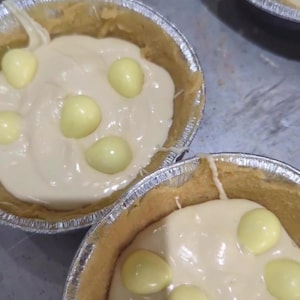 May include: Four round aluminum foil tart shells filled with a smooth, light beige cream and topped with small, pale yellow egg-shaped candies. A white rectangular label with black text reads Milky bar, positioned over the bottom tart.