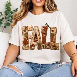 Könnte beinhalten: Ein cremefarbenes Kurzarm-T-Shirt mit dem Wort FALL in großen Buchstaben im Patchwork-Stil. Die Buchstaben sind mit herbstlichen Motiven verziert, darunter ein Fuchs, ein Vogel, Pilze, Eicheln, Blätter und verschiedene Knöpfe.