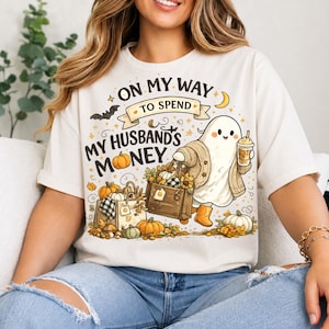 Könnte beinhalten: Ein cremefarbenes T-Shirt mit einer Grafik eines Geistes in Strickjacke und gelben Stiefeln, der einen Kaffeebecher und eine Einkaufstasche hält. Der Text ON MY WAY TO SPEND MY HUSBAND'S MONEY ist von Kürbissen, einer Fledermaus und Sternen umgeben.