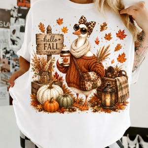 Könnte beinhalten: Ein weißes T-Shirt mit einer Grafik einer Gans, die eine Leoparden-Schleife, eine Sonnenbrille, einen Strickpullover und einen Schal trägt. Die Gans hält einen Kaffeebecher neben einem Holzschild mit der Aufschrift hello FALL. Kürbisse und Blätter.
