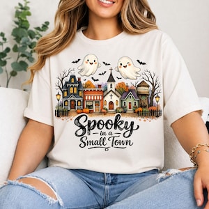 Puede incluir: Una camiseta de color crema con un gráfico de un pueblo pequeño con una iglesia, casas y una torre de agua. Dos fantasmas blancos y murciélagos negros vuelan sobre los edificios. El texto Spooky in a Small Town aparece en letras negras.